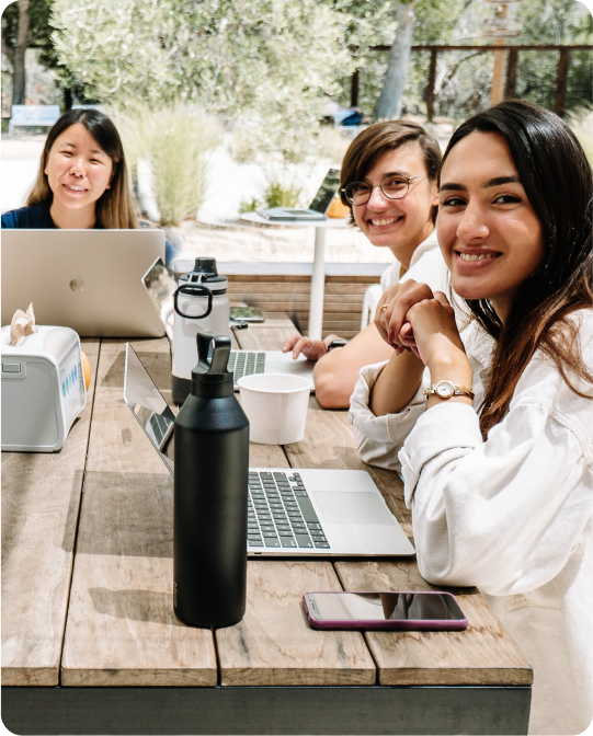 Three smiling people working on laptops at an outdoor wood table.