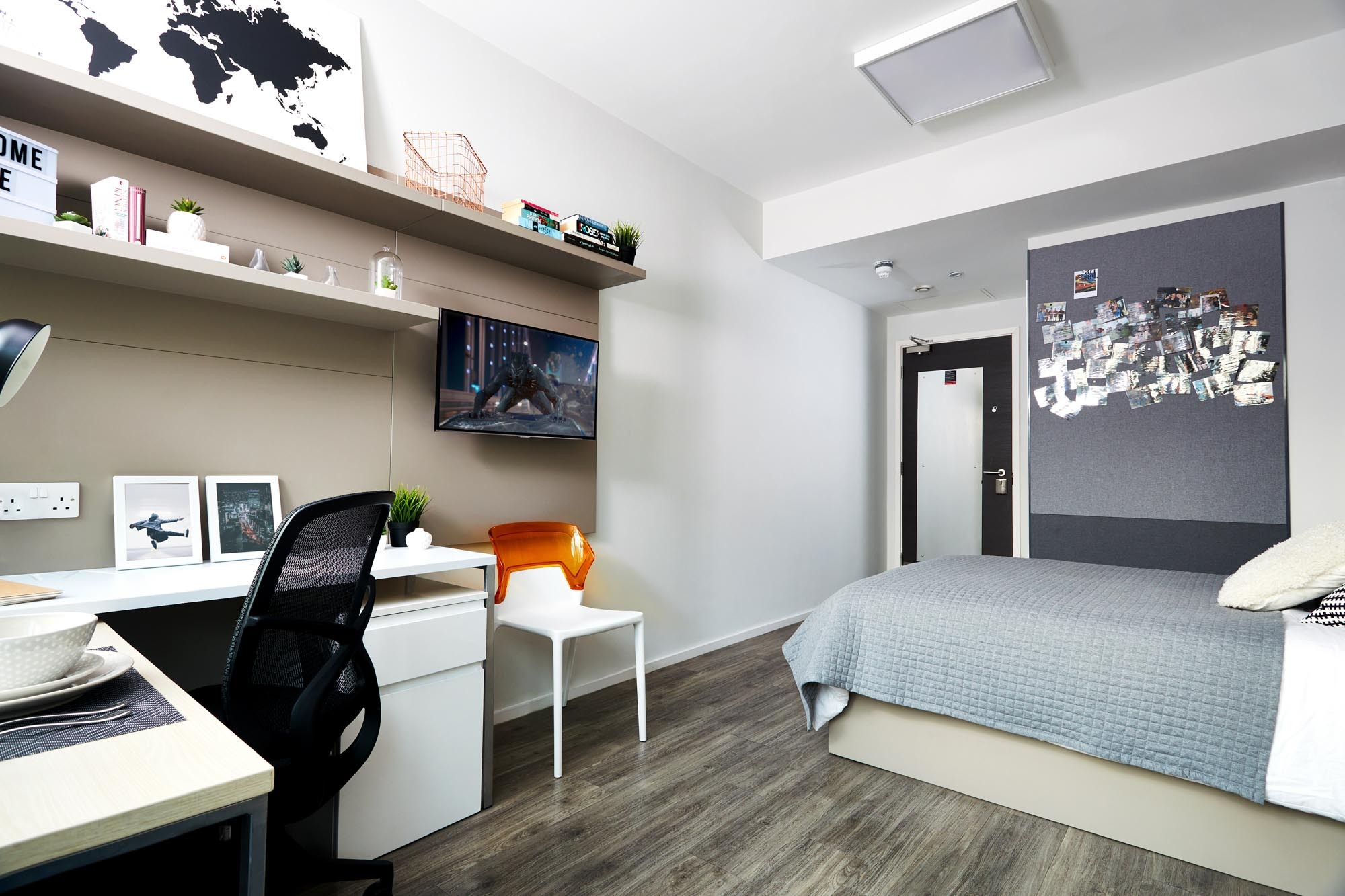 Modern dorm room with a bed, white desk, chair, and shelving. A wall-mounted TV displays an image. A bulletin board with photos is above the bed. The room has a light wood floor and a world map decor on the wall. A small lamp and potted plant are on the desk.