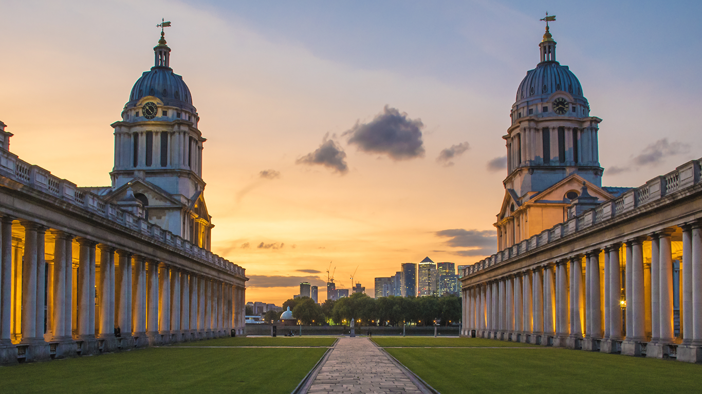 A symmetrical view of the Old Royal Naval College in Greenwich, London, during sunset captures the essence of life. The twin domed buildings frame a path with illuminated columns lining either side. The sky is painted with warm hues, and city skyscrapers are visible in the distance.