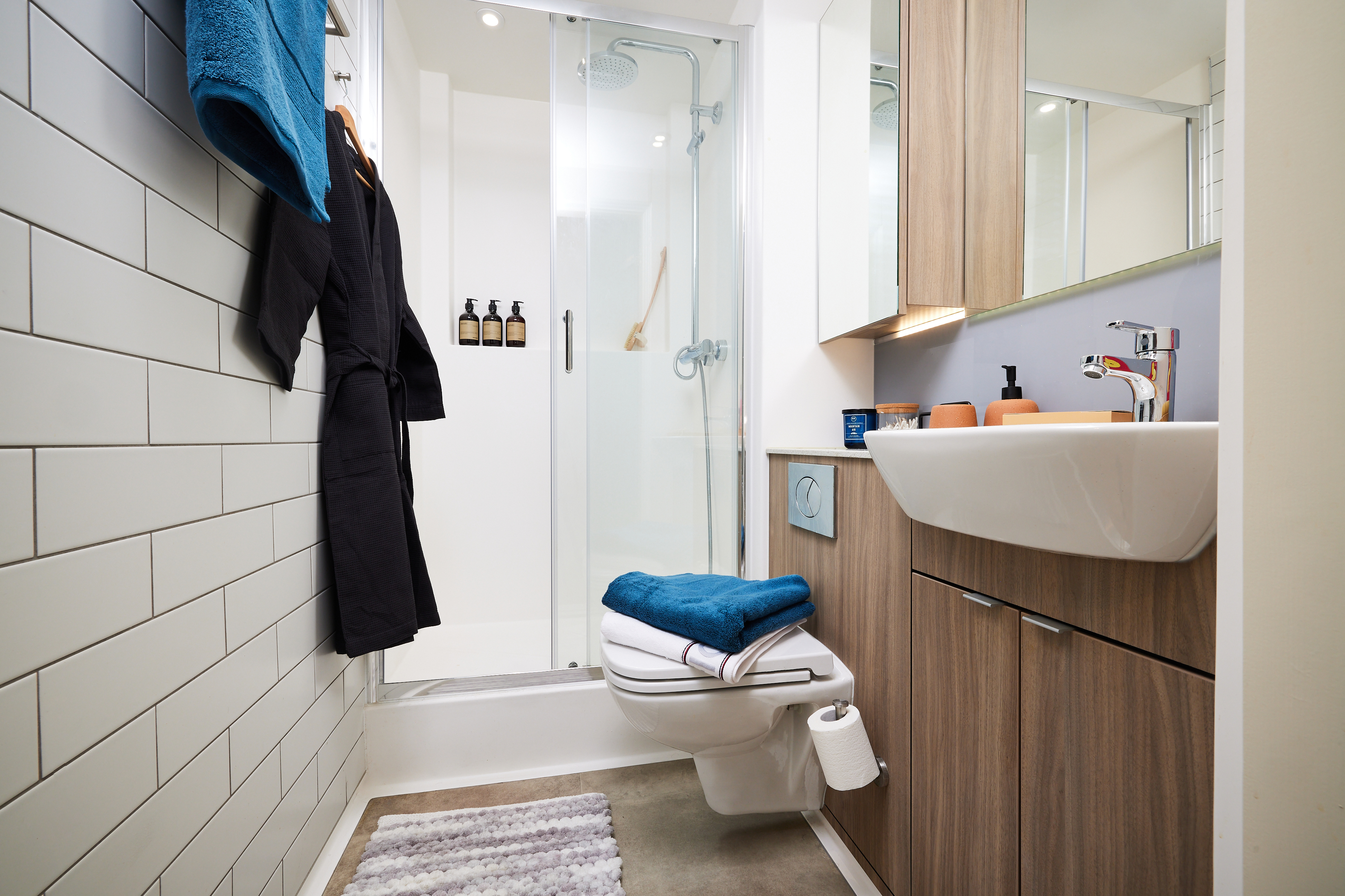 A modern bathroom with a glass-enclosed shower, white sink, and toilet. Wooden cabinets line the wall. Towels hang; one blue and one white are stacked on the toilet. There's a blue towel on a hook and several bottles in the shower area.