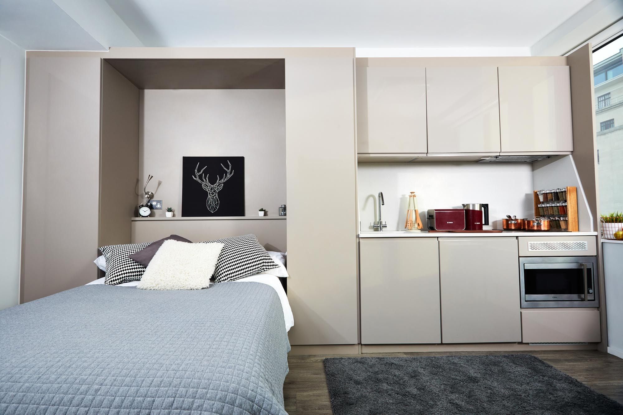 A modern studio apartment featuring a neatly made bed with gray and white bedding. Adjacent is a compact kitchen with sleek cabinets, a microwave, and a toaster. A deer silhouette artwork decorates the wall above the bed.