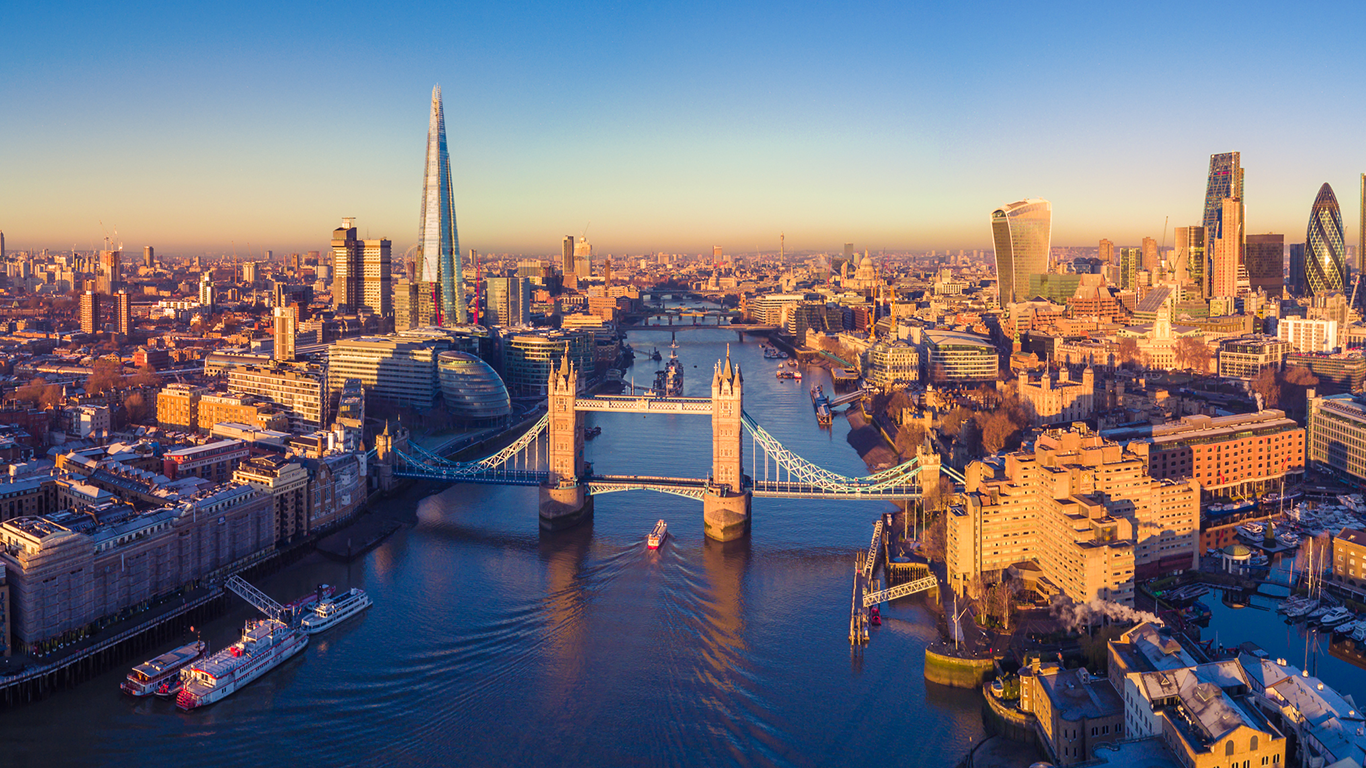 Aerial view of London at sunset, where vibrant city life unfolds along the Thames River. Tower Bridge stands proudly in the foreground, with The Shard punctuating the skyline. The clear sky casts a warm golden hue, illuminating the dynamic lifestyle of this iconic cityscape.