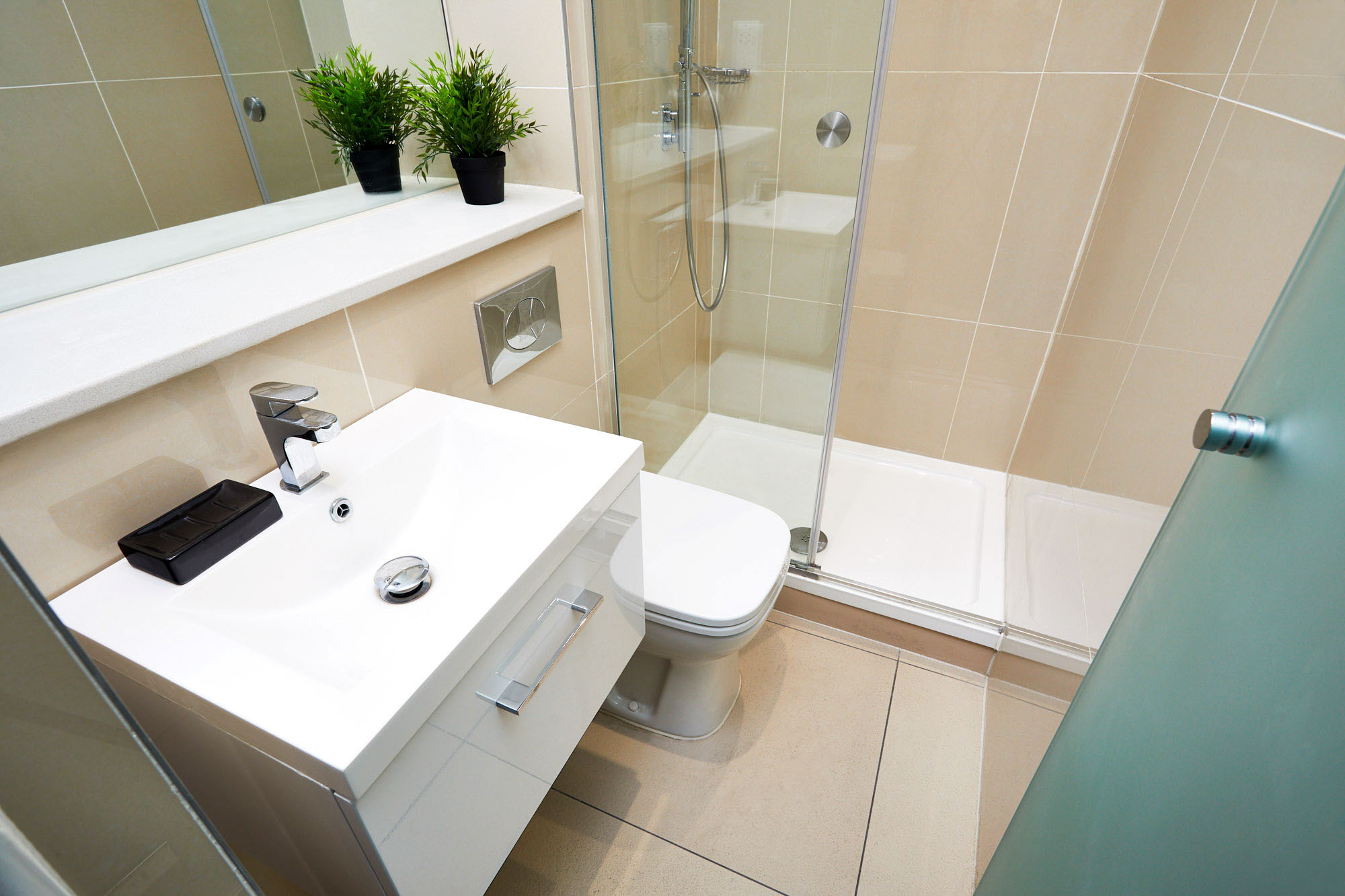 A modern bathroom with a white sink and rectangular mirror on the left, a toilet in the center, and a glass-enclosed shower on the right. Two small potted plants are on the countertop. Beige tiles cover the walls and floor.