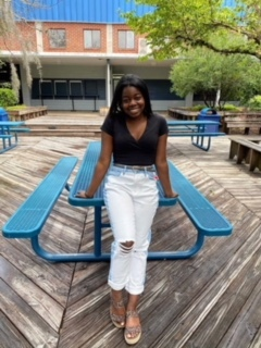 Young Black woman, Sheila Hodges, leaning against an aqua blue table and wearing a black top, white jeans with a belt, and sandals.