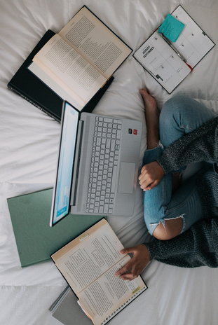 Woman in bed surrounded by laptop and books by Windows from unsplash