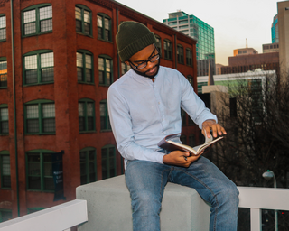 Black Poet Gabriel Angrand sitting, wearing glasses, in blue jeans, light-blue button-up, and green hat, holding a book in his hands.