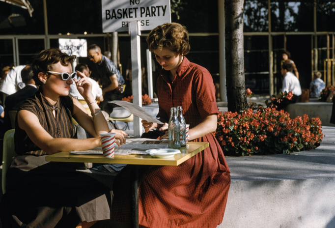 two women sitting at table outside with glasses and a newspaper