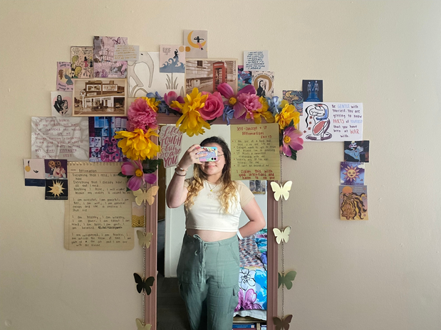 Girl taking selfie in wall mirror with art decorated around mirror.