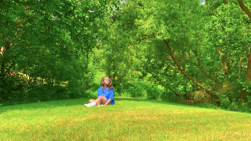 Girl is in a blue outfit, sitting in a field of green grass with trees in the back