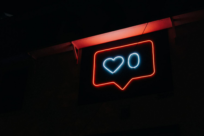 A neon light sign of a notification symbol. The inside contains a blue heart with the number zero next to it.
