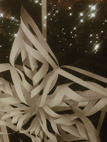 3-D paper snowflake shown against backdrop of a window with Christmas lights reflecting