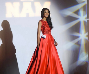 Girl is walking on stage smiling in a red gown with a trophy