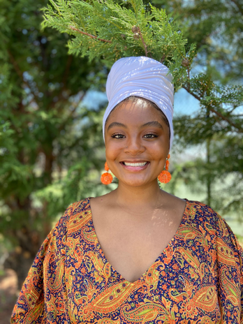 Girl is in a colorful dress and white headwrap smiling with trees in the background