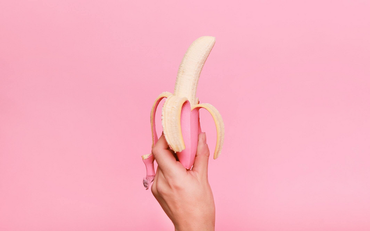 hand holding up a peeled banana in front of a pink background