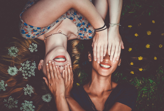 Two girls laying on a field, covering each other\'s eyes with each other\'s hands