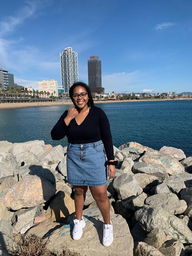 Young Black woman, Mercedes Owens, standing in front of the beach wearing a black top, jean skirt, and white shoes.