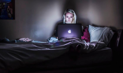 A young woman is sat in bed with a laptop in her lap. There is minimal lighting in the image except for the woman\'s face, which is illuminated by the laptop screen.