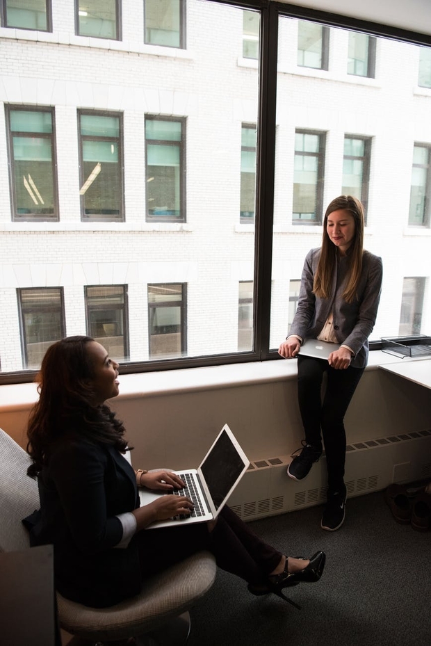 two women talking while holding laptops by the window at office