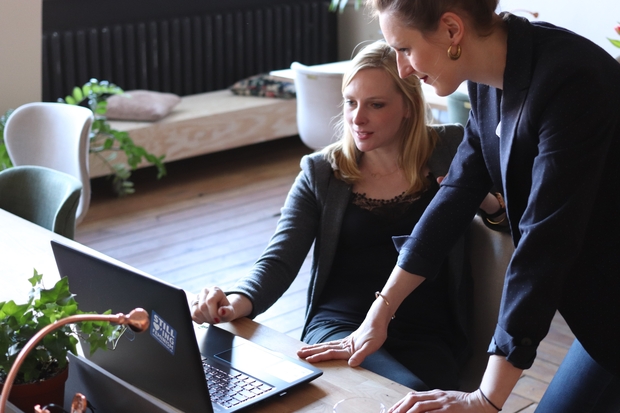 two women sit in front of a computer. there are plants on the desk. they are wearing blazers.