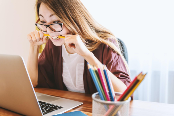 woman with glasses biting pencil while looking at laptop