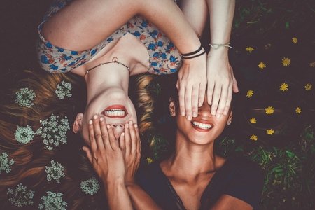 two woman smiling while laying on lawn field and covering each other's eyes