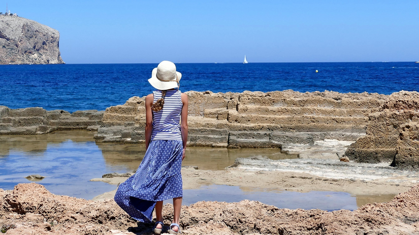 woman in blue dress and white hat facing the ocean