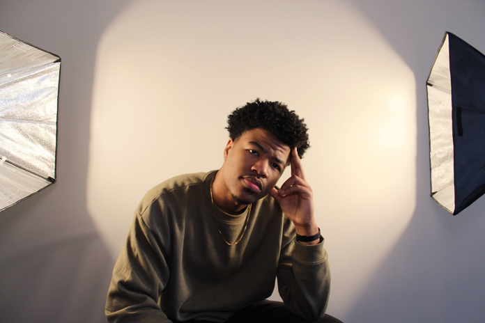 Young Black artist NiSPLASH sitting on a chair between 2 lights, in front of a white backdrop.