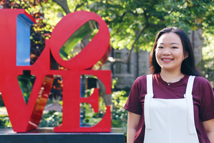 Young Asian woman, Jessica Bao, standing in front of the LOVE statue wearing a maroon shirt with white overalls.