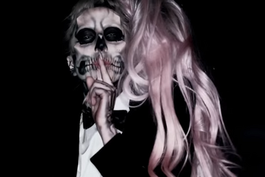 Lady Gaga in a skull makeup, pink hair and indicator finger on her mouth