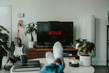 Anonymous individual holds onto a remote, which is directed towards the TV screen (displaying the typical Netflix slogan), on what seems to be his/her living-room/bedroom.