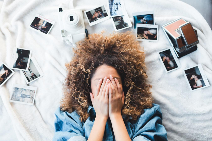 A young girl lies in bed with her hands covering her face, seemingly in distress. Her head is surrounded by printed photos and a camera.