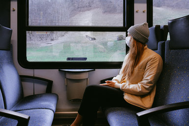 Woman sitting on a train with a beanie and mask on looking out the window.