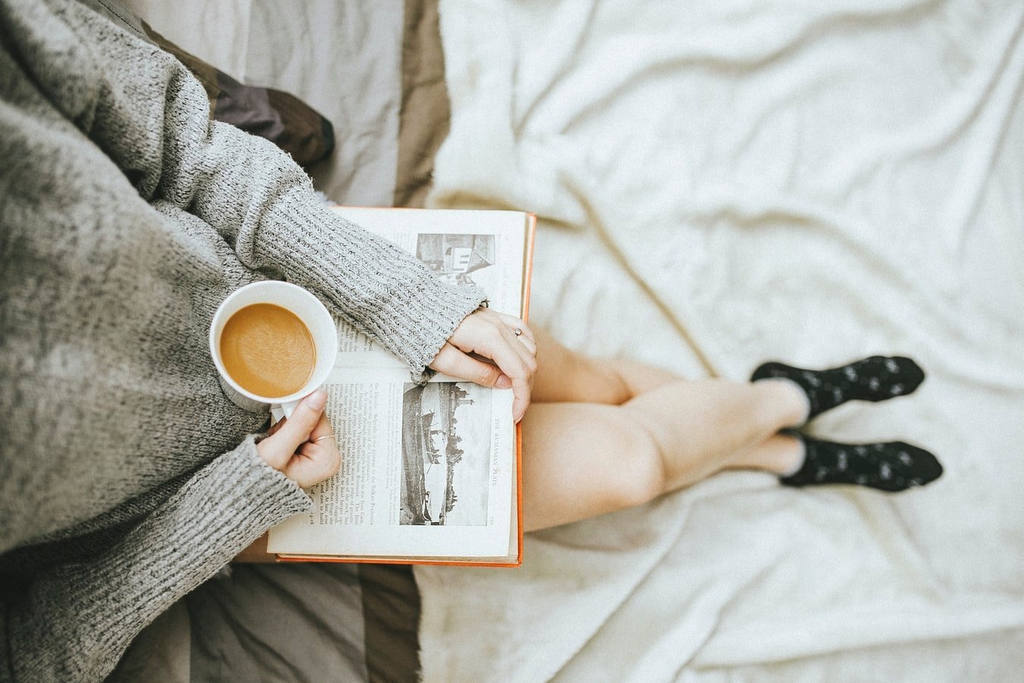 woman holding cup of coffee and reading a book while sitting on bed